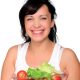 Eine Schwangere dunkelhaarige Frau lächelt und hält eine Glasschüssel mit Gemüse und Salat.