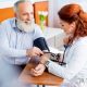 Ein Mann mit grauem Bart sitzt lächelnd einer rothaarigen Ärztin im weißen Kittel gegenüber. Sie hat das Blutdruckmessgerät an seinem linken Oberarm angelegt und fühlt gleichzeitig mit ihrer linken Hand seinen Puls.