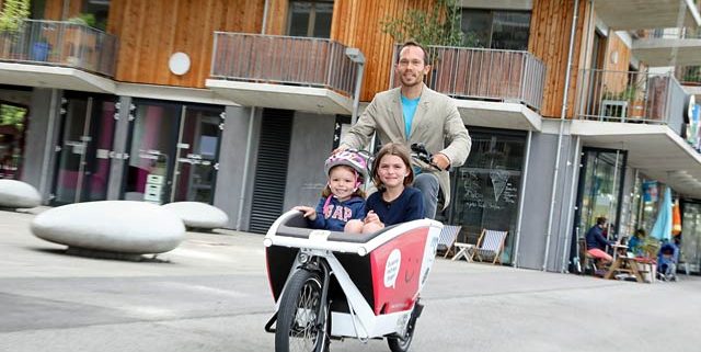 Ein Mann fährt mit einem Lastenrad, in dem 2 Kinder sitzen
