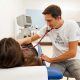 Ein Mann mit weißen T-shirt mit der Aufschrift „Neunerhaus“ hört das Herz einer auf einer Krankenliege liegenden Person mit einem Stethoskop ab.