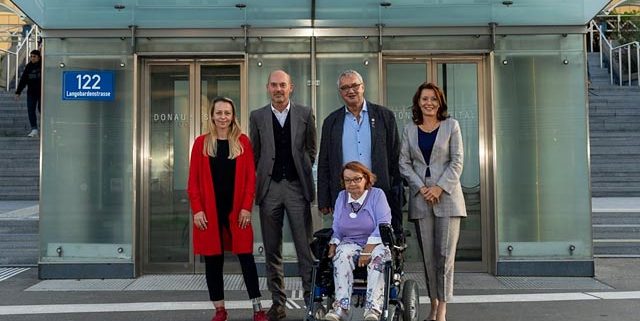 Drei Frauen, davon eine sitzend im Rollstuhl, eine mit Beinprotese, und zwei Männer befinden sich vor dem Zugang mit Lift zum Donauspital