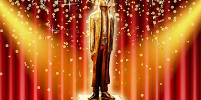 Eine Schwarze Statue eines Mann mit einem Hut und langen Mantels steht auf einem goldenem Podest vor einem roten Vorhang. Sie ist von zwei Scheinwerfern beschienen und es regnet Konfetti.