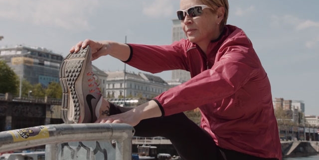 Eine Frau in Jogging-Bekleidung steht im Freien an einem Fluss in der Stadt und dehnt die Beinrückseite an einem Geländer.