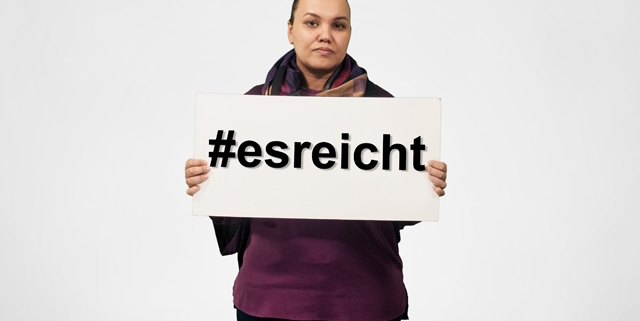 Eine Frau mit ernstem Blick steht vor einem weißen Hintergrund und hält ein Schild vor der Brust mit dem Text "#esreicht".