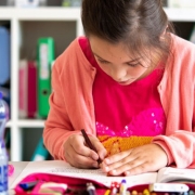 Ein Mädchen malt auf der Schulbank ein Heft an, im Vordergrund steht eine Wasserflasche.